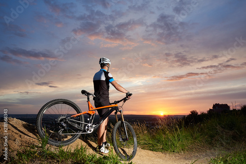 Back view of cyclist male in a helmet and sports clothes rests on the background of a sunset from a height watching the mountain in the distance, the enchanting sky and the bright sun