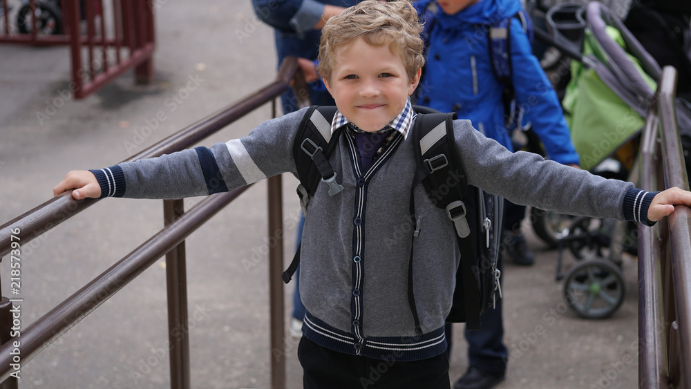 Little smiling Caucasian curly boy in school uniform with backpack comes up on stairs.
