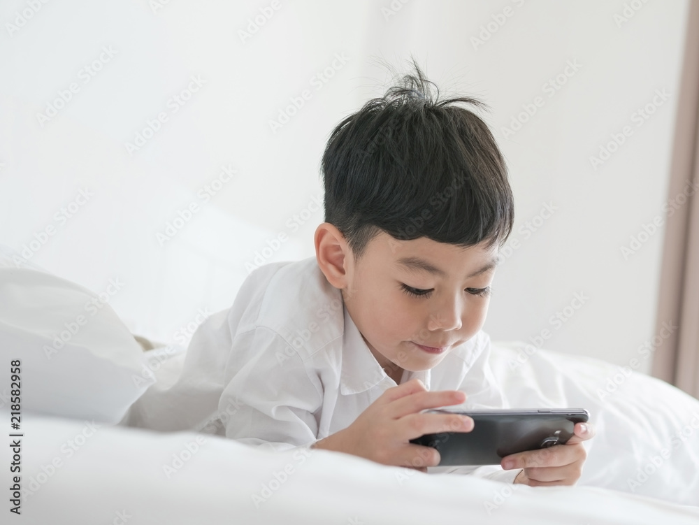 Cute little asian kid focused on smartphone while lying on the bed. Boy playing games on smartphone. Social and technology concept Cute little asian kid focused on smartphone while lying on the bed. Boy playing games on smartphone. Social and technology concept