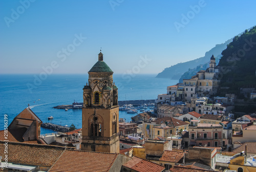 A stunning view of Amalfi town from above, with high ancient bell tower in the middle and mountains covered with greenery in the background. Shot before sunset during the golden hour