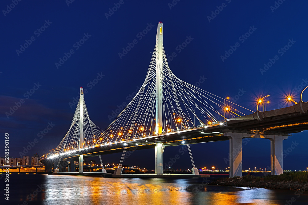 Obraz premium The bridge of circle highway road over Neva river near the mouth of it in the blue hour after the sunset. Night view on the buildings of Petersburg city and the Finish gulf