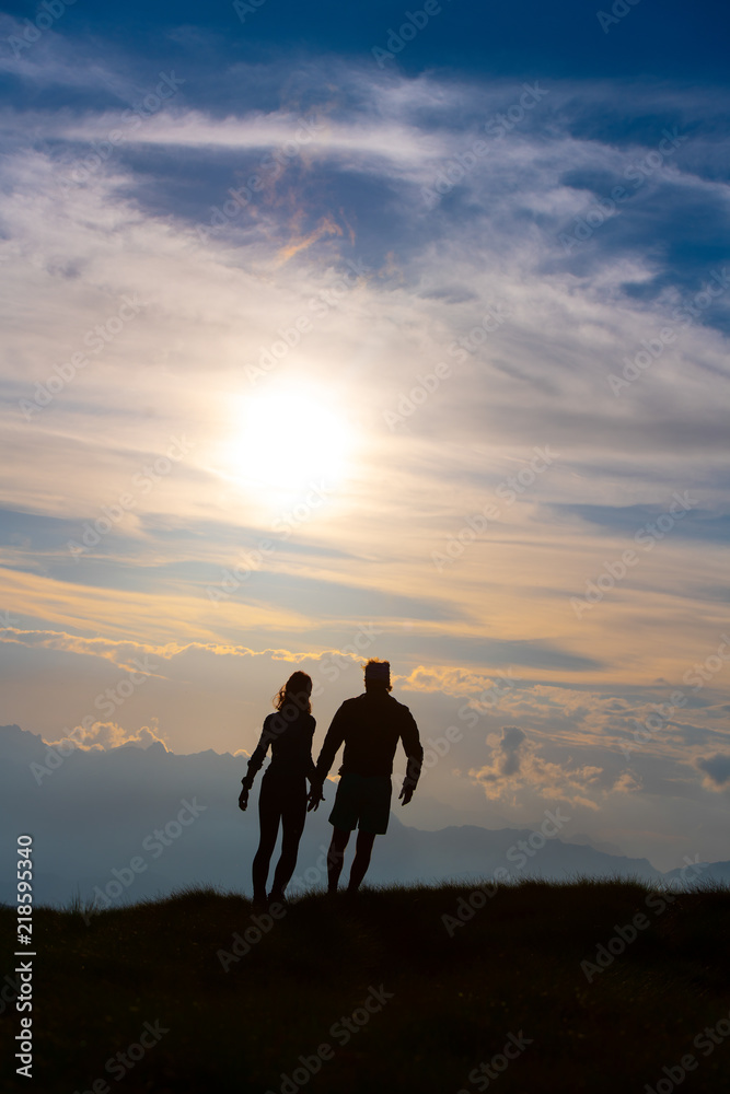 Couple in silhouette holding hands walking towards colorful clouds at sunset in the mountains