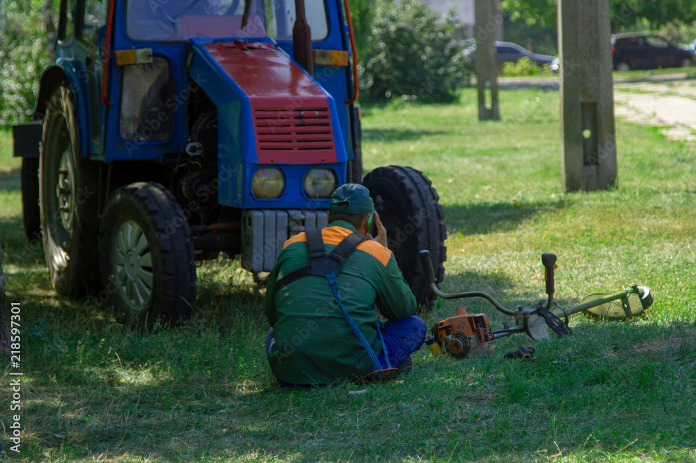 Obraz premium Lazy worker rests after cutting the lawn. A city utility worker is sitting on the grass and talking on the phone. The worker idles during working hours near the tractor.