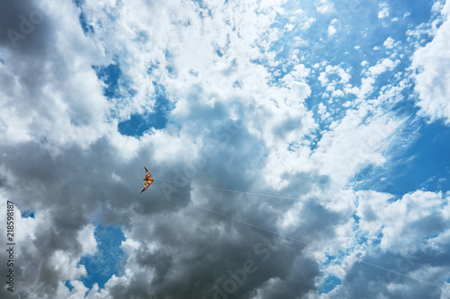 A large kite floats high in the sky among the clouds against the blue sky and the bright summer sun and it is controlled by two white ropes going to the ground