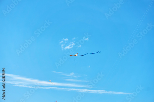 A large kite floats high in the sky among the clouds against the blue sky and the bright summer sun and it is controlled by two white ropes going to the ground