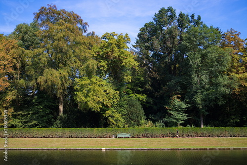 Side view of man jogging forward  green bench on the street parallel the border of the poor with background of range high tree and bush at Schlosspark Benrath, Düsseldorf, Germany 