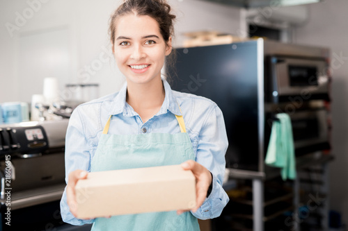 Waist up  portrait of smiling young  woman wearing apron holding box with takeaway food and looking at camera, copy space