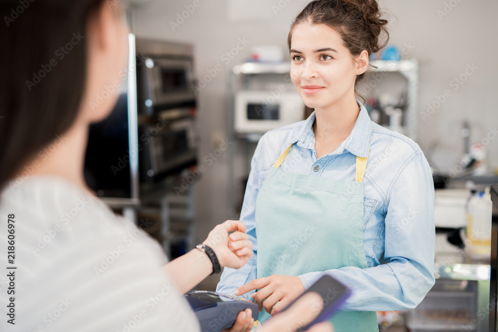 Obraz premium Waist up of smiling young woman wearing apron looking at customer paying via NFC in shop, copy space