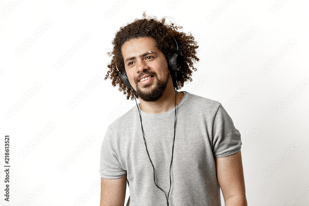 A curly-headed handsome man wearing a gray T-shirt and ripped jeans is standing and listening to music in the headphones with a soft smile over the white background. A curly-headed handsome man wearing a gray T-shirt and ripped jeans is standing and listening to music in the headphones with a soft smile over the white background.