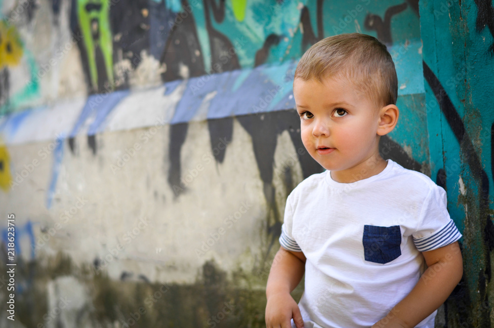 Fototapeta premium A two-year-old baby boy in hip hop style clothes sits under a wall with green graffiti and looks inquiringly upwards