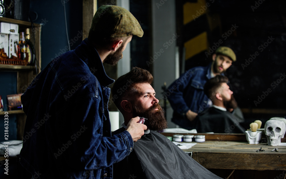 Barber busy with grooming beard of hipster client, mirror reflexion on background. Grooming concept. Hipster with beard covered with cape trimming by professional barber in stylish barbershop