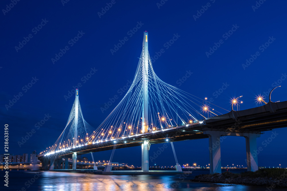 Obraz premium The bridge of circle highway road over Neva river near the mouth of it in the blue hour after the sunset. Night view on the buildings of Petersburg city and the Finish gulf