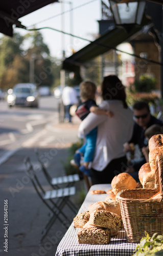 Fresh tasty bread is on a table in a street next to bakery. In the background: family with small children is in a street cafe, a car drives along a city road, people walk along a sidewalk.