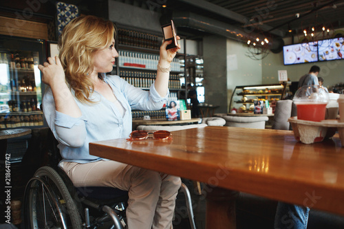 Woman in a wheelchair is drinking coffee and making selfie on a smartphone in a cafe.