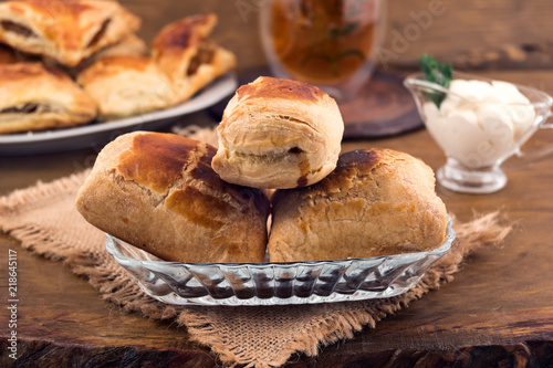 Sweet dessert-puff pastry with cheese filling in a plate on a linen napkin, gravy boat with sour cream, a glass of herbal tea, all on a wooden background.