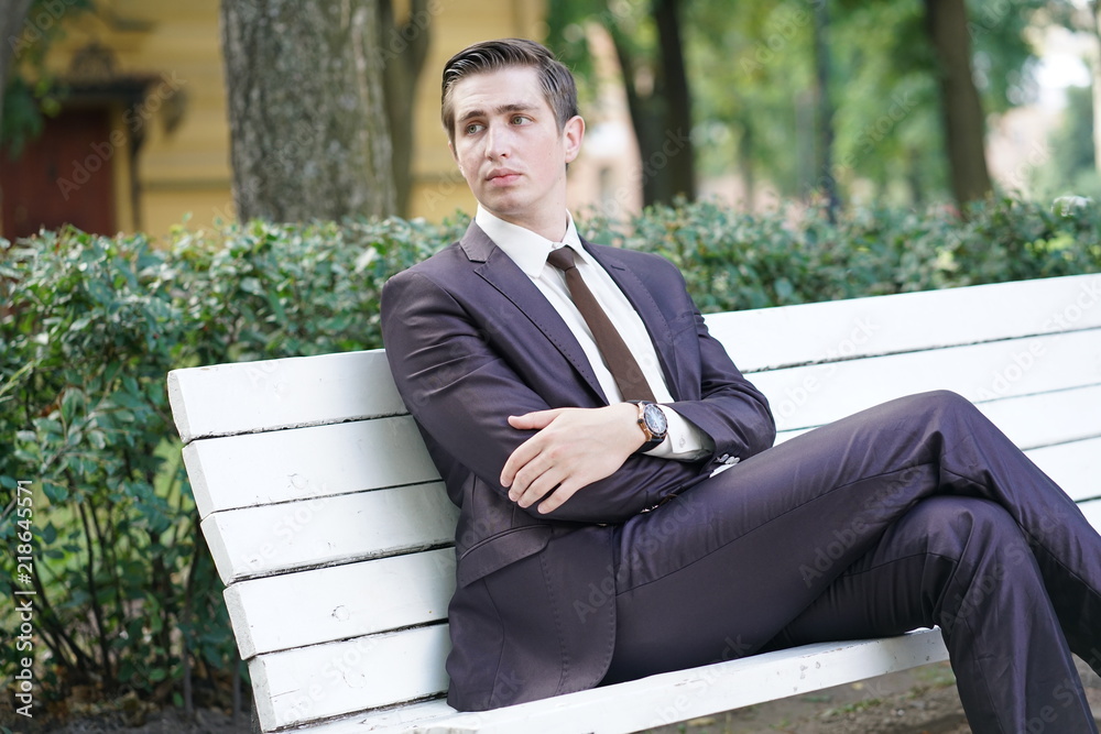 A tired man in a business suit left the office and went in the Park. he is sitting on a white bench alone and wait someone. A tired man in a business suit left the office and went in the Park. he is sitting on a white bench alone and wait someone.