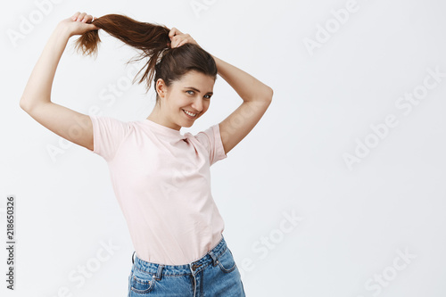 Studio shot of carefree and emotive cute european girlfriend combing long brown hair making hairstyle while pulling strands with hands and smiling broadly at camera liking her haircut © Cookie Studio