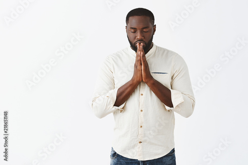 Indoor shot of focused and determined very serious African American man in white shirt, closing eyes, holding hands in pray and making wish, praying or hoping for better over gray background