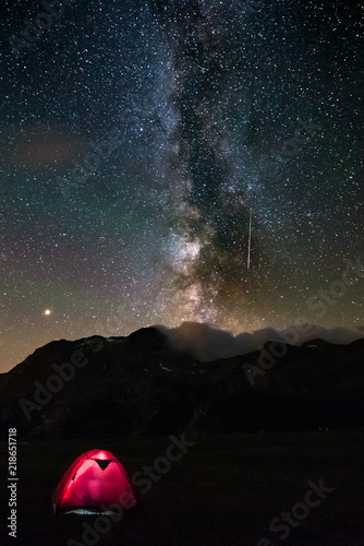 Meteor with green tail starry sky milky way. Illuminated tent in the foreground, camping on the Alps. Mars Planet on the left. Adventure and exploration in summertime.