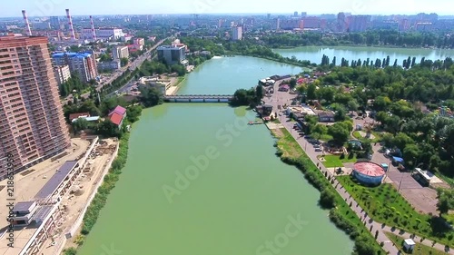 Aerial footage over city Central Park view from top attractions of Kuban river, Krasnodar summer green trees bridge, multi houses skyline visible in distance. Descend green park and birds flying away.