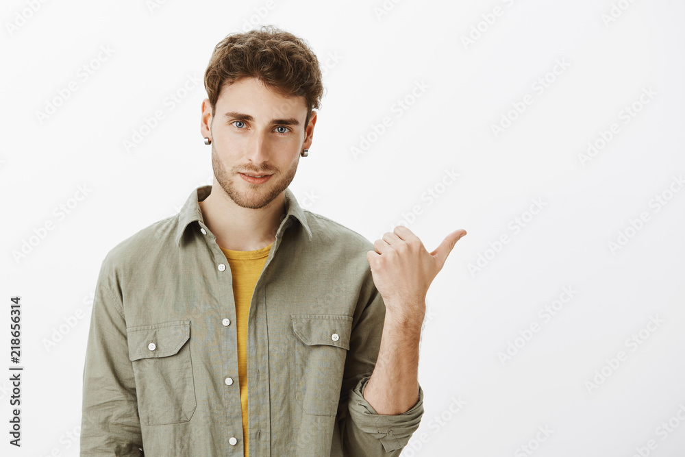 Man pointing at friend intoducing him to girlfriend. Handsome confident young male with curly hair, pointing behind or right with thumb and gazing at camera, waiting for answear on question