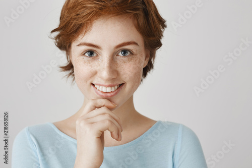 Eyes are mirror of soul. Close-up portrait of attractive female with ginger hair and freckles holding hand on chin and smiling broadly, gazing at camera with interested expression over gray wall