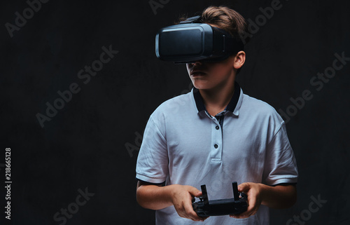 Portrait of a young boy dressed in a white t-shirt using virtual reality glasses and holds a control remote. Isolated on a dark background.