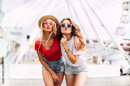 Best friend having fun near Ferris wheel, going crazy together, wearing hats and mirrored sunglasses, amazing view on the city, bright colors evening sunlight