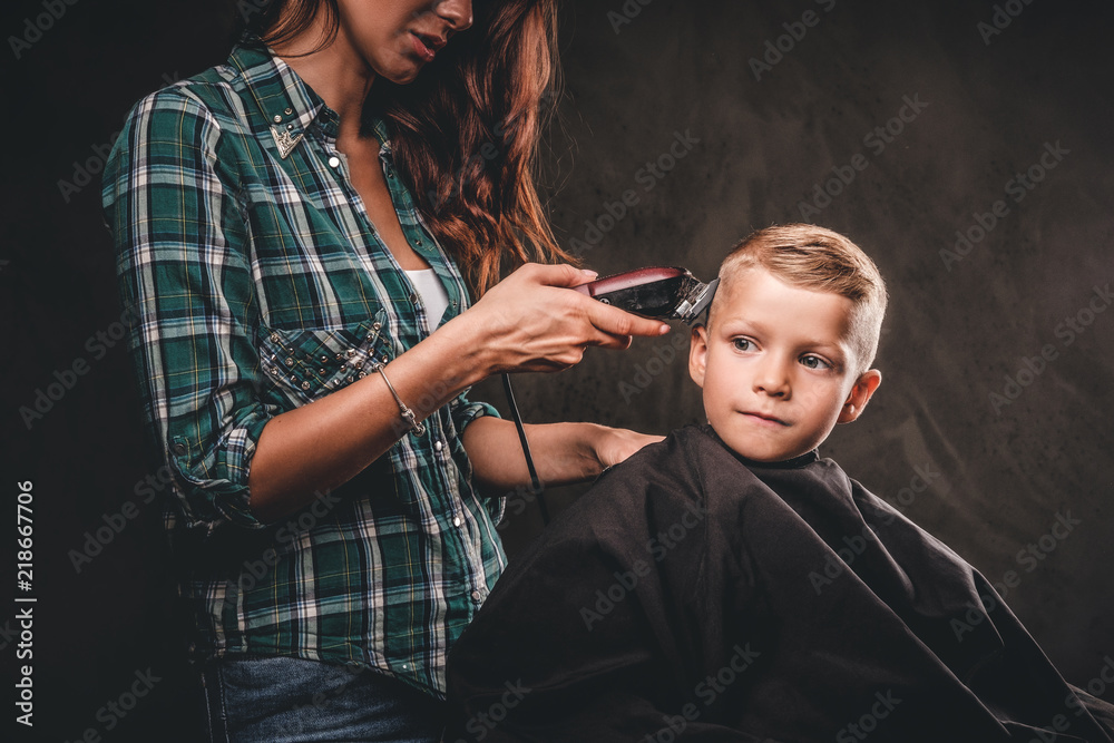 Obraz premium Children hairdresser with the trimmer is cutting little boy against the dark background. Cute preschooler boy getting haircut. 