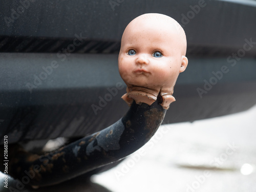 Tablou pe pânză Doll's head impaled in irreverent manner to car tow-bar, with bumper of car in b