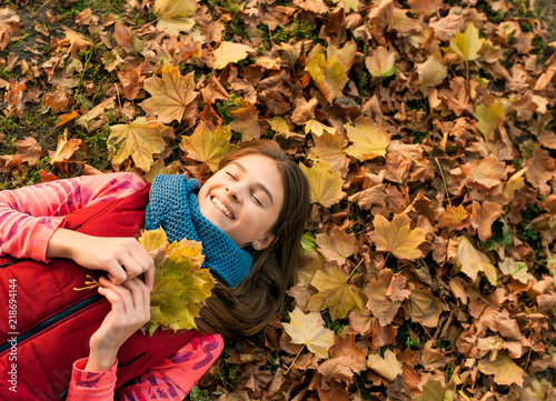 Smiling young girl lying down on autumn maple leaves at fall outdoors. Portrait of a beautiful smiling teenage. Young teenager girl looking to camera. Autumn  winter season.
