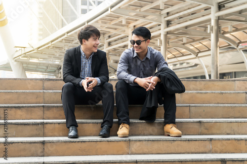 Happy two young businessman sit on the stairs talking. Attractive businessmen sitting on stair at outdoor in city after work together talking about fun.