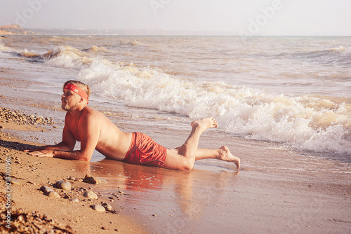 guy in red swimming trunks and a bandana on his head, lies on a sandy beach and is covered by a beautiful foaming wave. The concept of rest, lifestyle and good mood.
