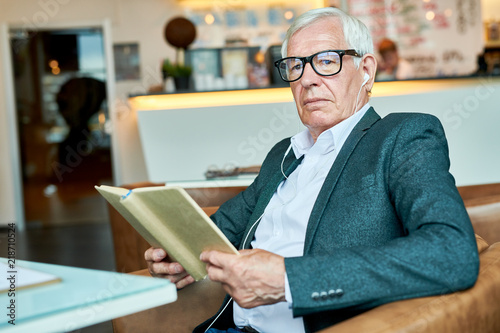 Portrait of successful senior businessman reading book in cafe while relaxing during coffee break and looking away pensively