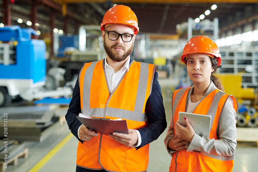 Waist up portrait of two modern factory workers wearing hardhats and reflective vests posing in production workshop, holding clipboard, copy space Waist up portrait of two modern factory workers wearing hardhats and reflective vests posing in production workshop, holding clipboard, copy space