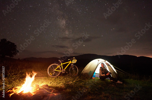 Young woman cyclist resting at night camping near burning campfire, illuminated tourist tent, mountain bike under beautiful evening sky full of stars. Outdoor activity and tourism concept