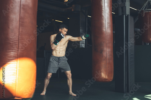 Shredded man is boxing with punching bags inside. He is hitting equipment with cross kick. Combat workout for strength and endurance concept