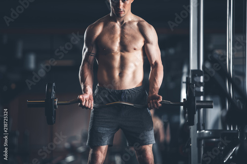 Bodybuilder is straining all body while making effort during workout. He is training upper body with weight. Male is standing and lifting barbell for biceps