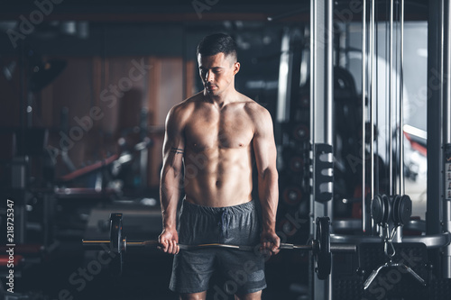 Shredded athlete is using barbell during workout in sport center. He is standing and lifting outfit before himself. Guy is exercising biceps muscles