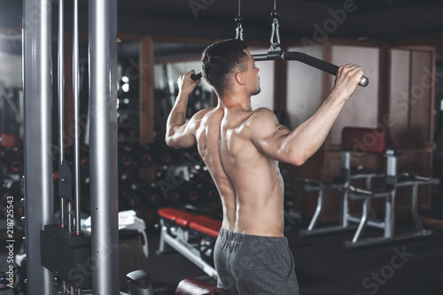 Shredded shirtless guy is exercising latissimus with sport equipment. He is standing and pulling down handles of vertical outfit. Man is working on dorsal relief