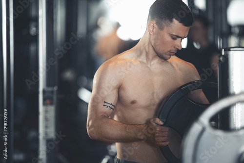 Topless tattooed man is using barbell for training strength and relief. He is checking equipment before workout while standing next to it. Male leading healthy lifestyle concept