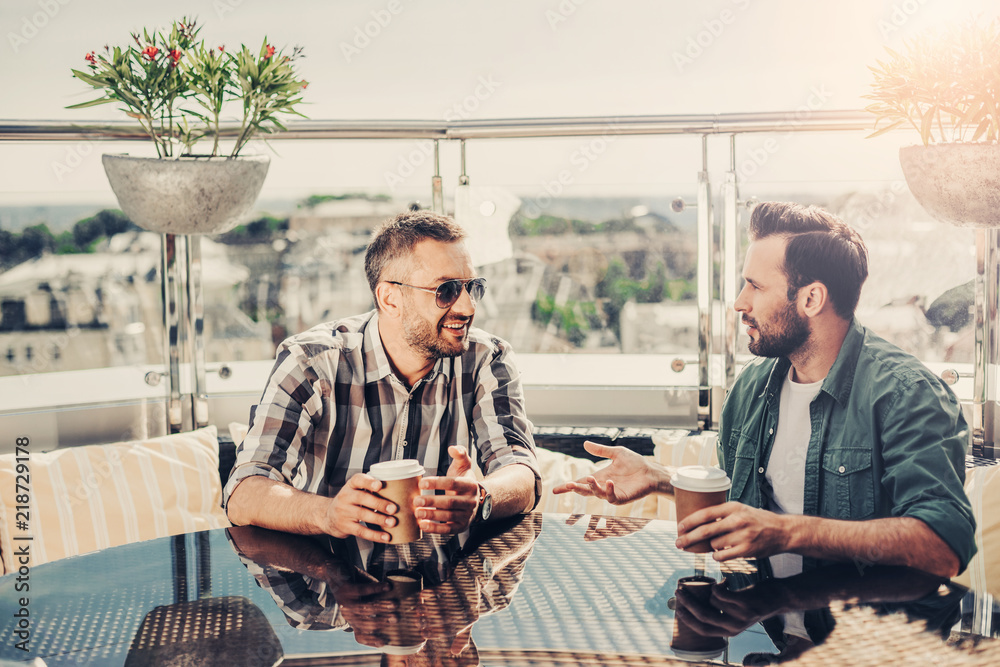 Interesting meeting. Male friends sitting at the table and communicating. They looking at each other and holding cups of coffee