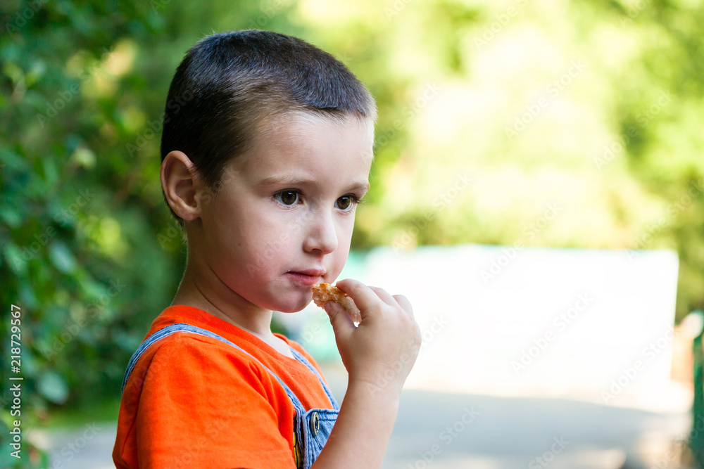A cute little boy who eats a piece of bread and looks away in a green city park on a summer warm day A cute little boy who eats a piece of bread and looks away in a green city park on a summer warm day