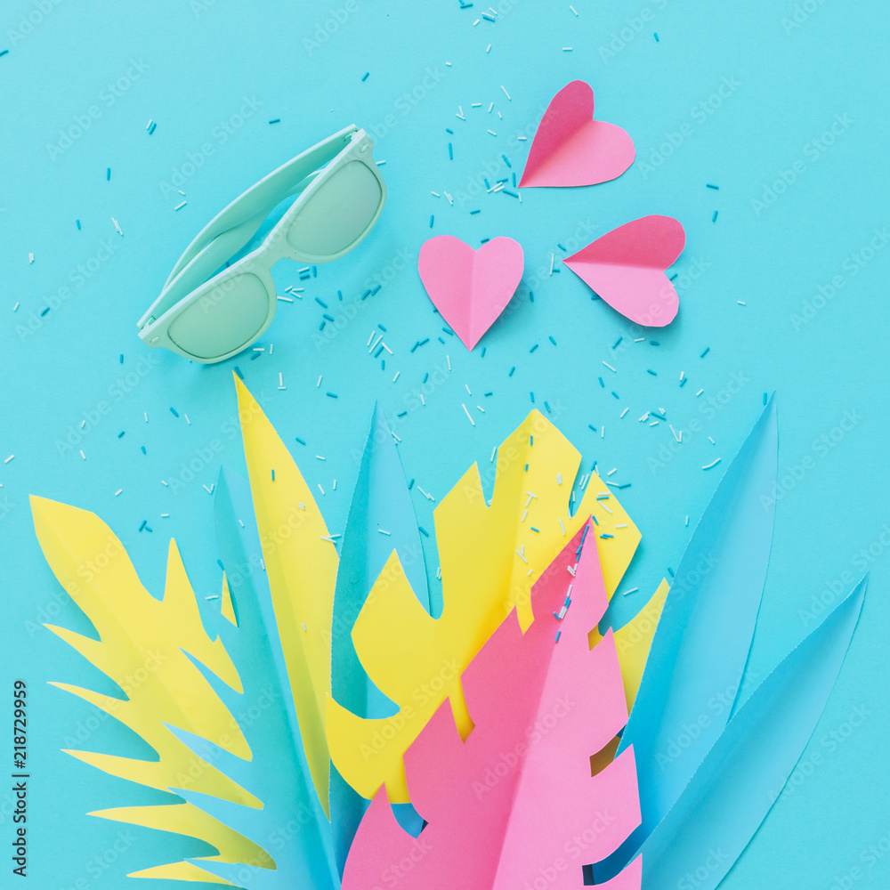Blue sunglasses and Pink hearts lie among tropical palm leaves made of paper