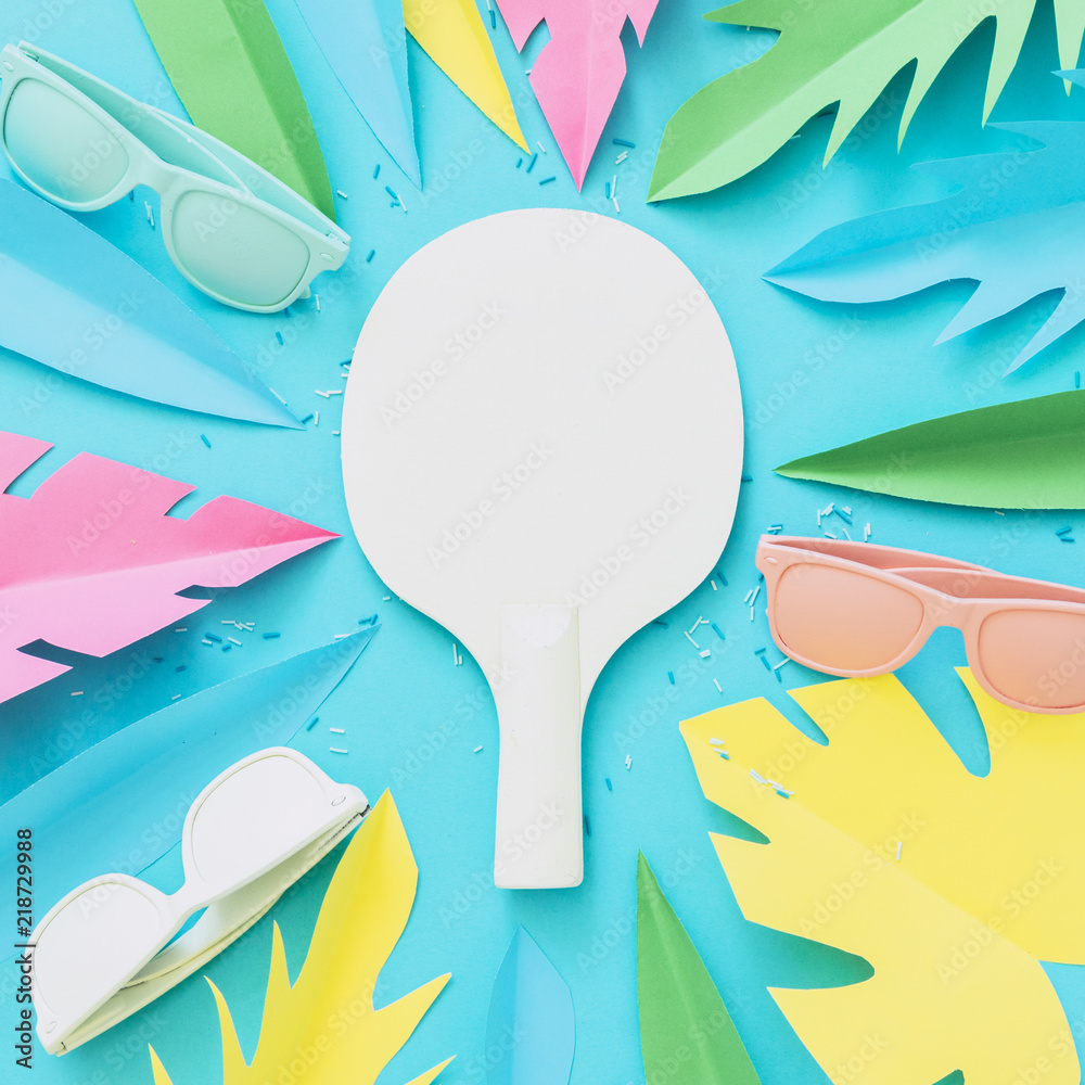 A tennis racket painted white lies among the tropical leaves of a palm tree made of paper and sunglasses. Summer concept of beach sports and recreation