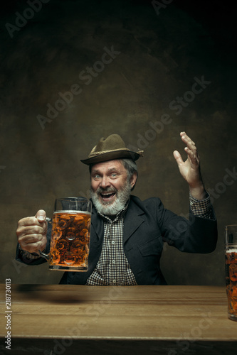 Smiling bearded man clinking with mug of alcohol in pub. Enjoying my favorite beer. The front view of handsome smiling senior man with glass of beer sitting at the wooden table. Studio shot with