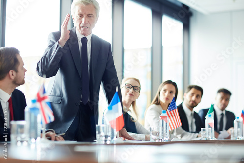 Aged confident politician in suit standing by table and debating with his foreign colleagues at political event photo