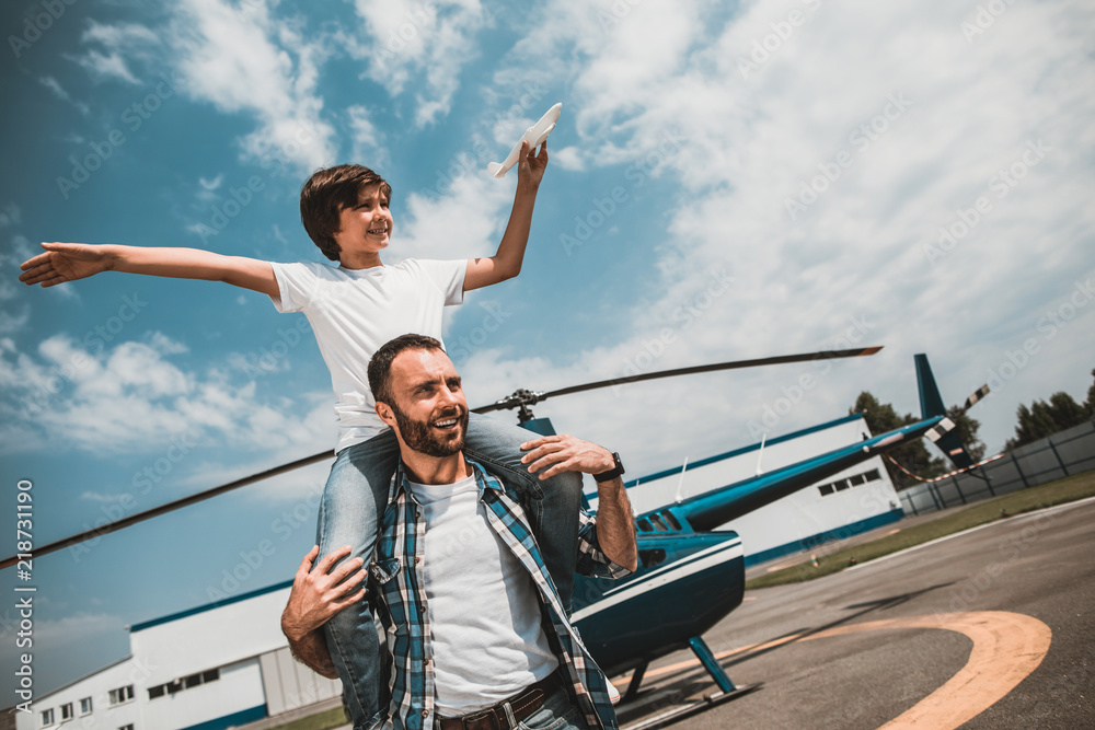 Portrait of satisfied boy playing with little plane while situating on shoulders of cheerful dad. They speaking together during game while situating opposite helicopter on air-stop Portrait of satisfied boy playing with little plane while situating on shoulders of cheerful dad. They speaking together during game while situating opposite helicopter on air-stop