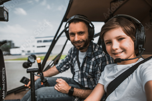 Portrait of satisfied unshaven man and cheerful boy wearing contemporary earphones while looking at camera. They having leisure in rotor plane as pilots