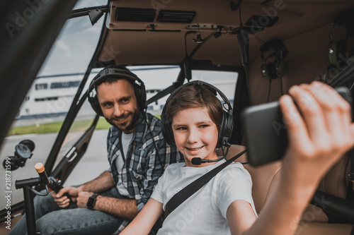 Portrait of happy bearded father and optimistic child taking photo on cellphone while wearing headsets. They spending time in helicopter © Yakobchuk Olena